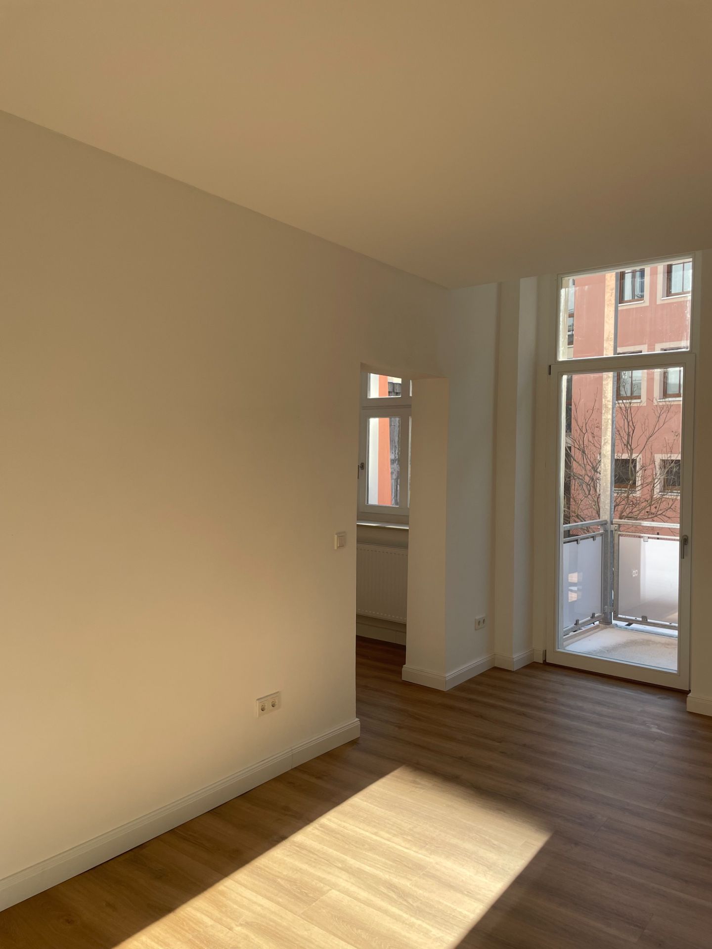 Heller Raum mit Holzfußboden, weißen Wänden und bodentiefen Fenstern zu einem Balkon.