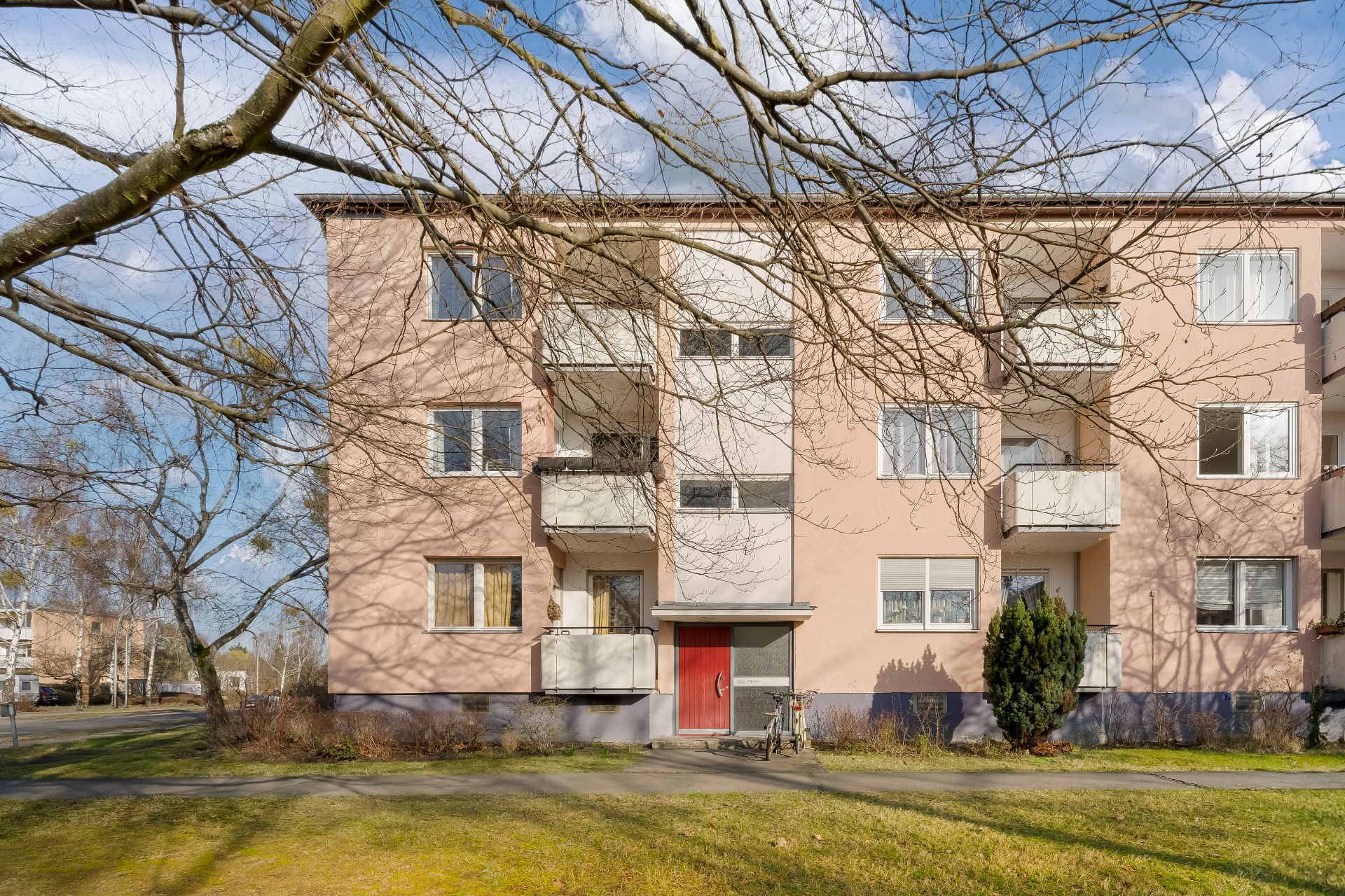 Mehrfamilienhaus mit Balkonen, roter Eingangstür und Fahrrad vor dem Gebäude.
