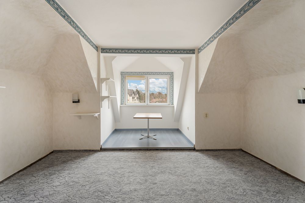 Heller Raum mit Fenster, kleinem Tisch, Regal und grauem Teppichboden.