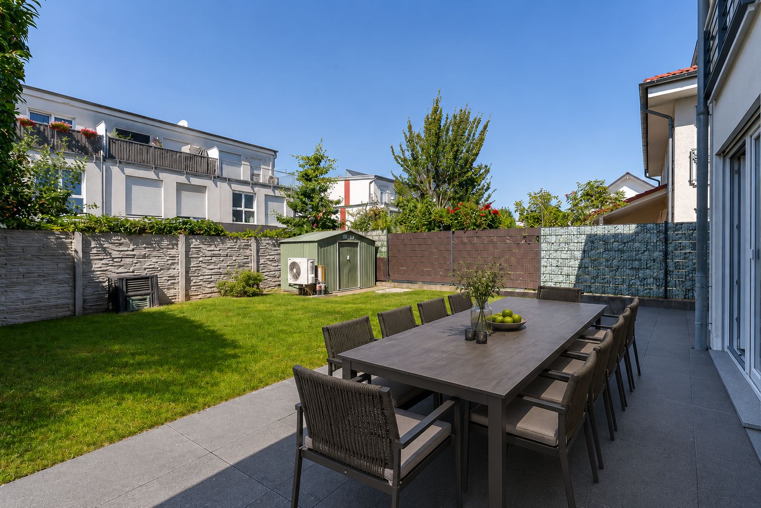 Garten mit großem Esstisch und Stühlen auf einer Terrasse, Rasenfläche und kleinem Geräteschuppen.