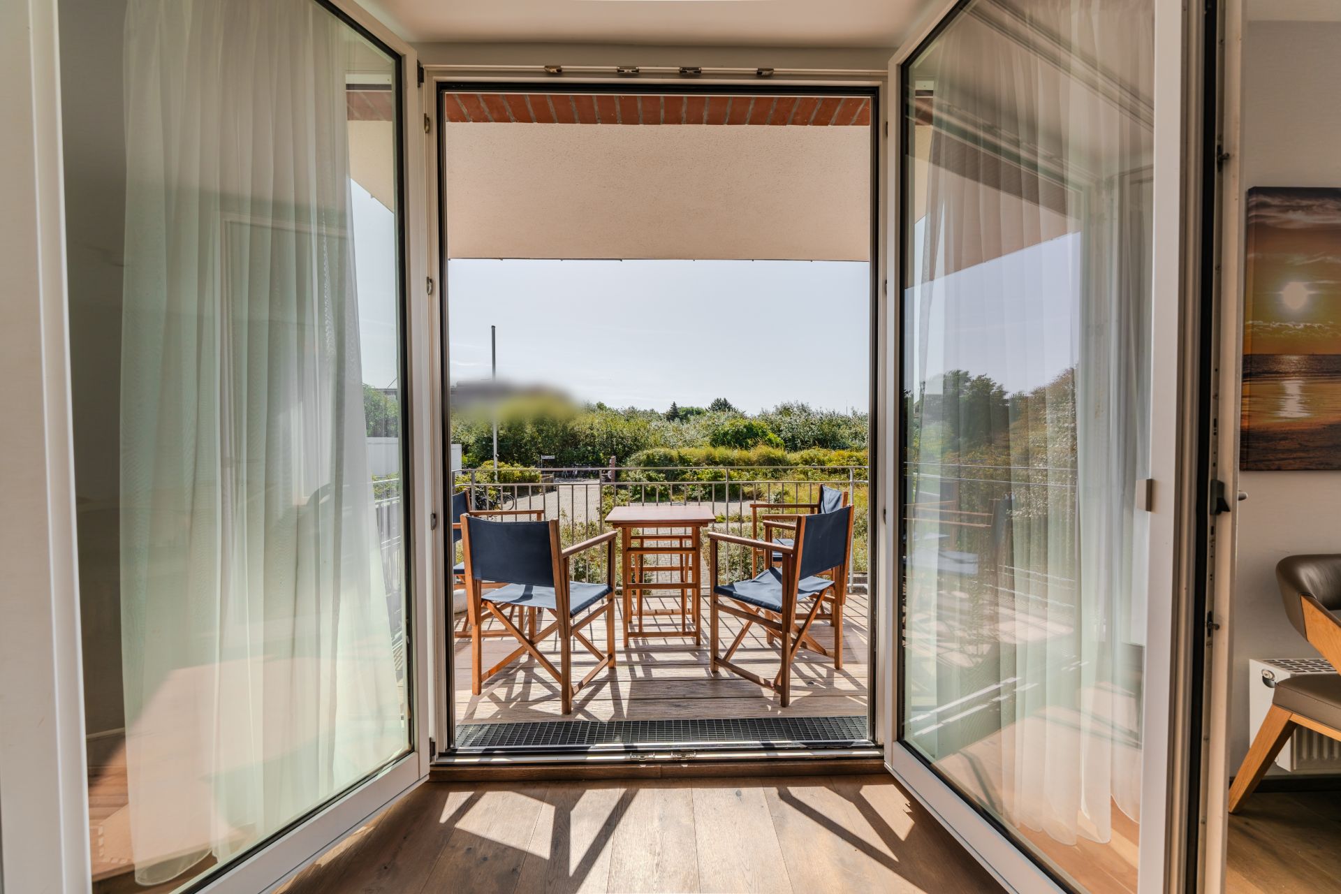 Offene Terrassentür mit Blick auf einen Balkon mit Holztisch und vier Stühlen, grüne Aussicht.
