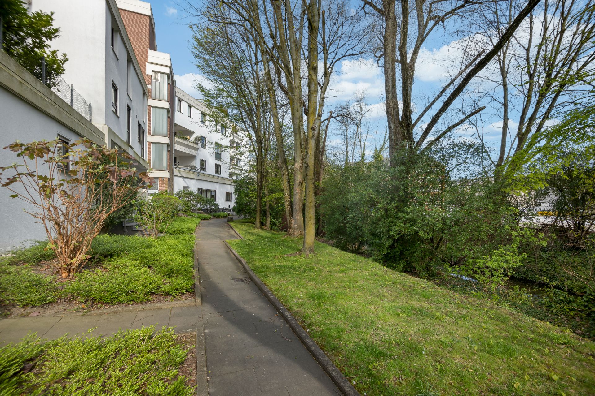 Gepflasterter Gehweg neben Wohnhaus mit Bäumen und grüner Bepflanzung.