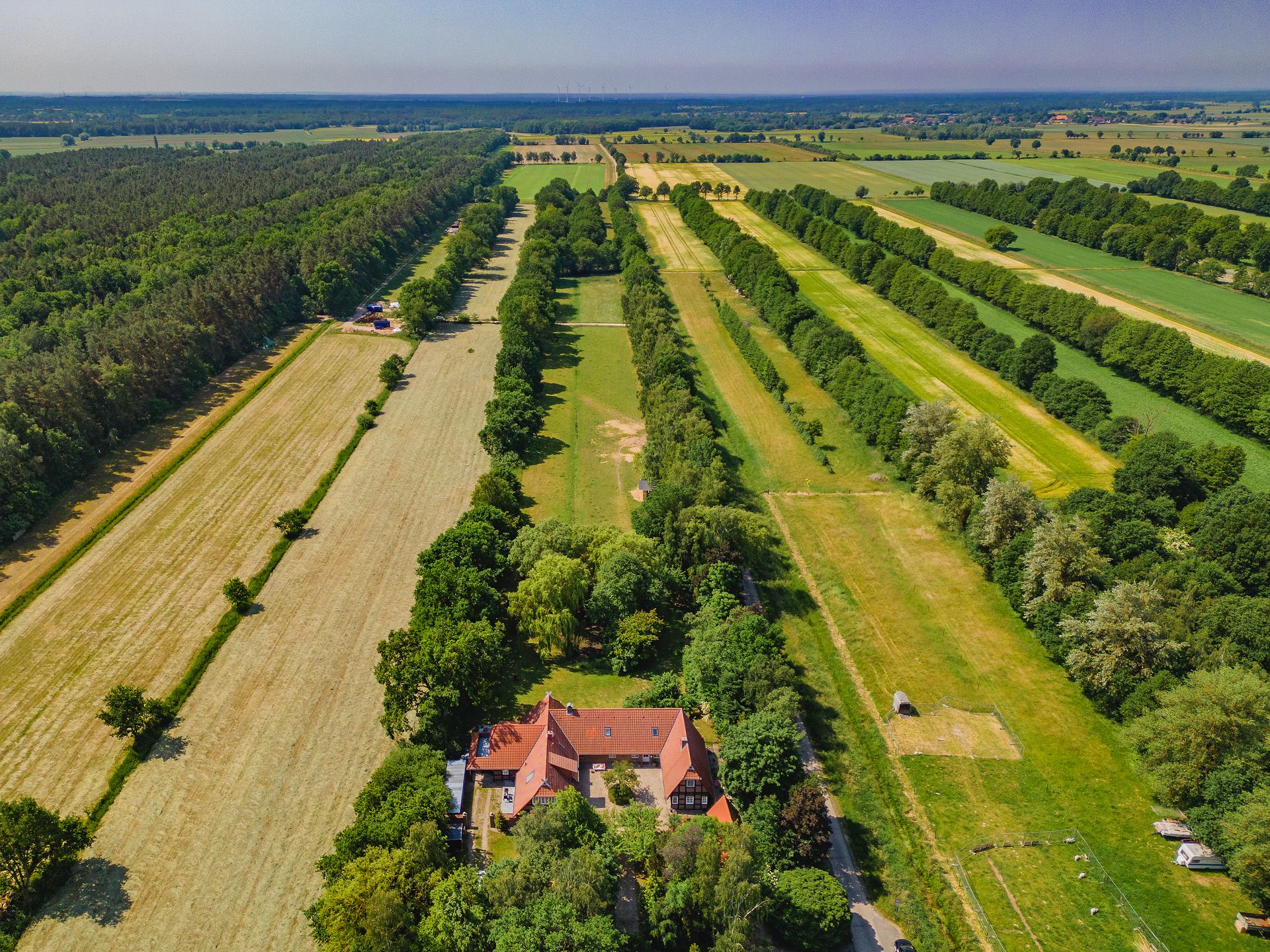 Luftaufnahme eines Hauses mit rotem Dach, umgeben von Wiesen, Feldern und Baumreihen.