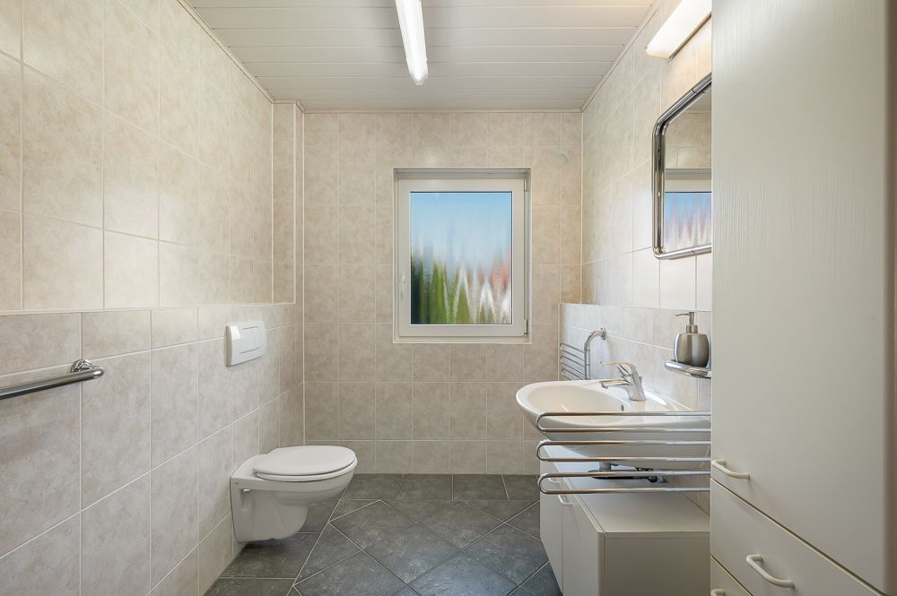 Helles Badezimmer mit Wandtoilette, Fenster, Waschbecken und Spiegel.