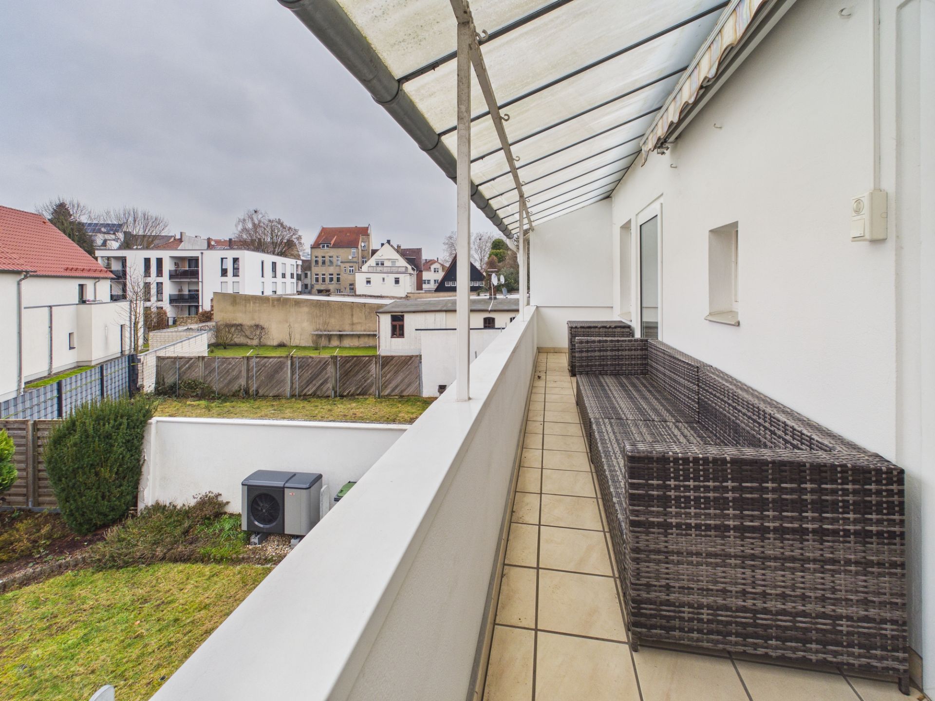 Überdachter Balkon mit gefliestem Boden und Rattan-Sofa, Blick auf Garten und Nachbarhäuser.