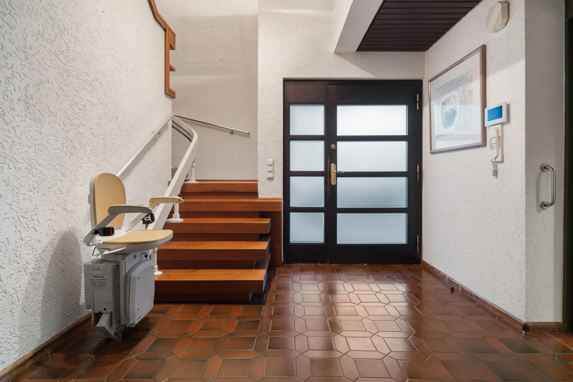 Hausflur mit Treppenlift, braunen Fliesen, Treppe und Glastür.