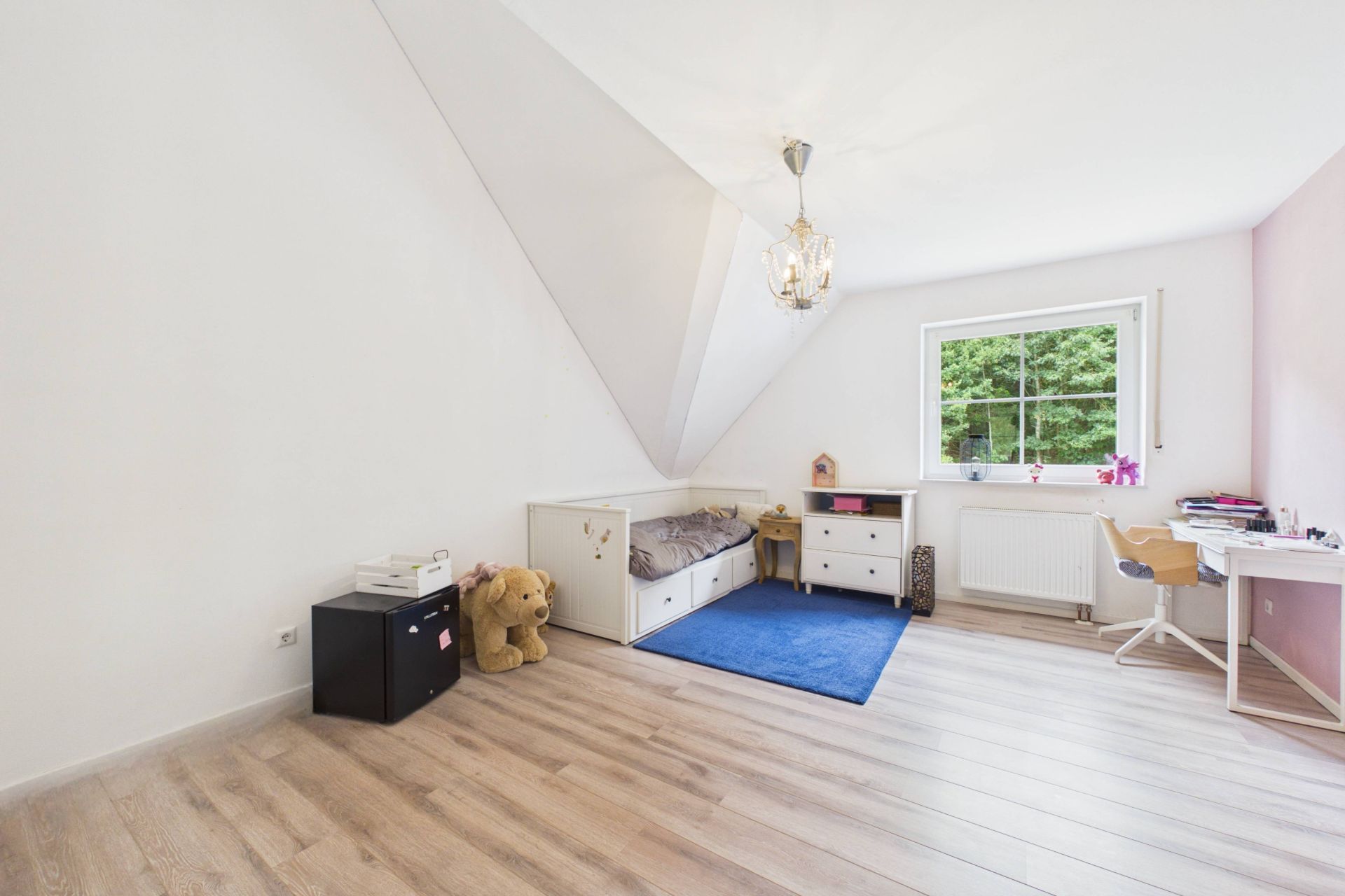 Helles Kinderzimmer mit Schreibtisch, Bett, Regalen, Kuscheltier und Teppich unter einem Dachschrägenfenster.