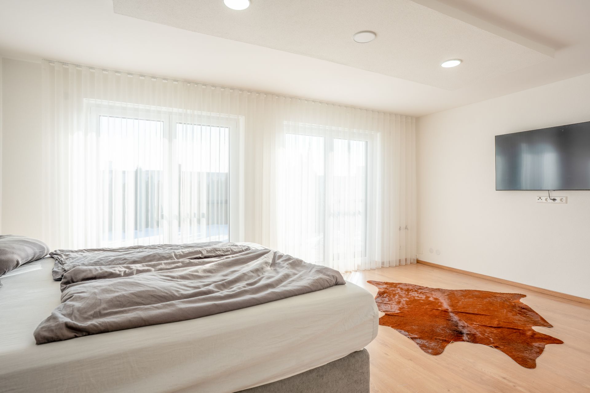 Helles Schlafzimmer mit Doppelbett, großem Fenster, Fernseher an der Wand und braunem Teppich auf Holzboden.