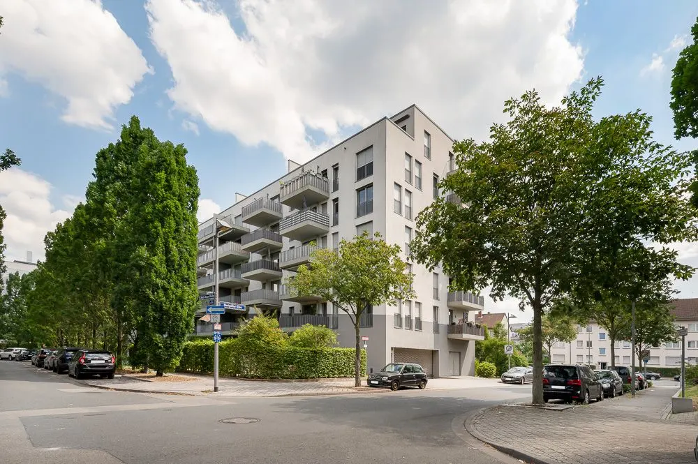Mehrstöckiges Wohngebäude an einer Straßenecke mit Balkonen und Bäumen davor.