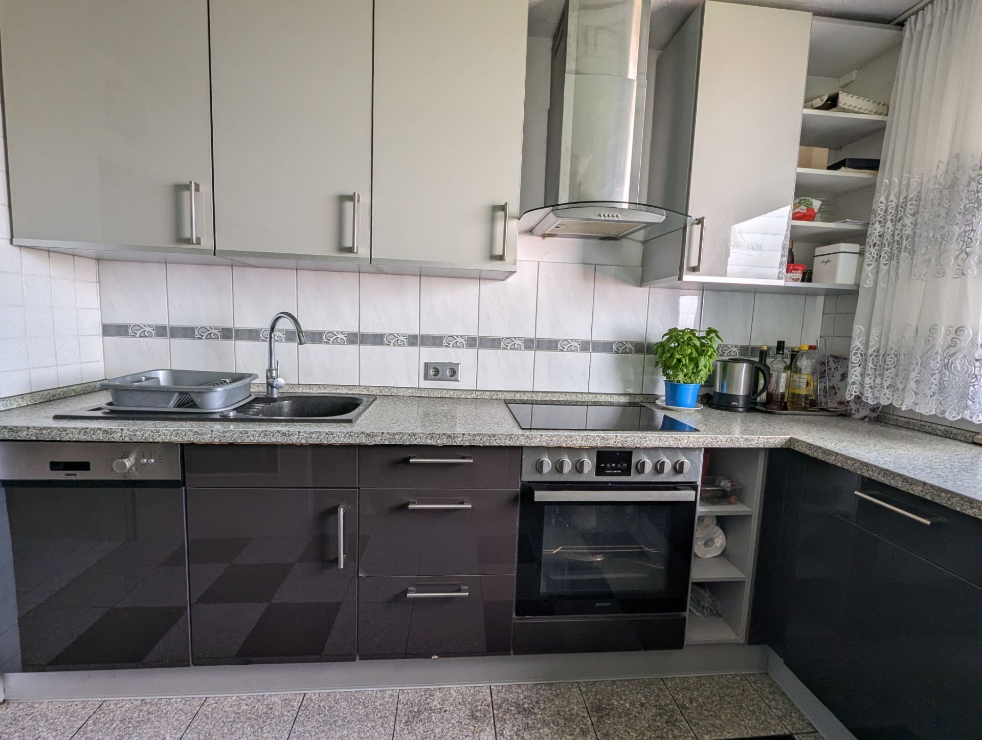 Moderne Einbauküche mit Herd, Spüle, Abzugshaube und Arbeitsplatte, daneben ein Fenster mit Gardine.