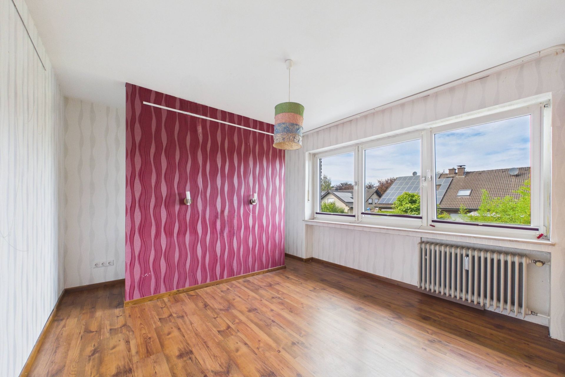 Leeres Zimmer mit großem Fenster, Holzfußboden und gemusterter rot-weißer Wand.