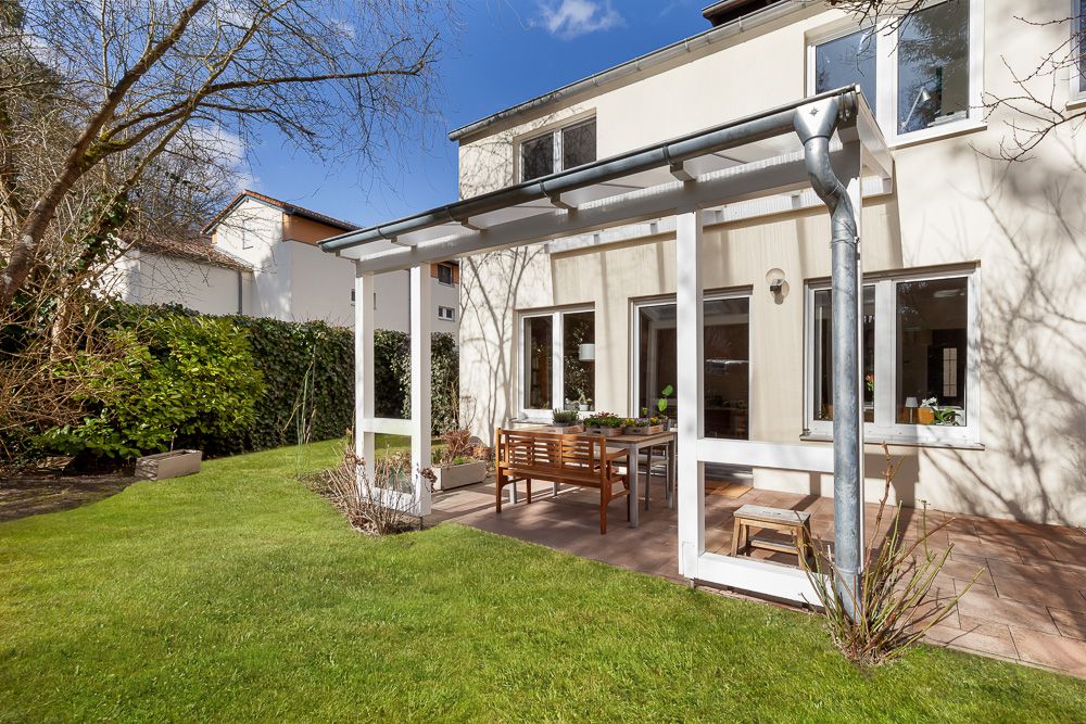 Überdachte Terrasse mit Holztisch und Stühlen, angrenzend an ein helles Wohnhaus im Garten.