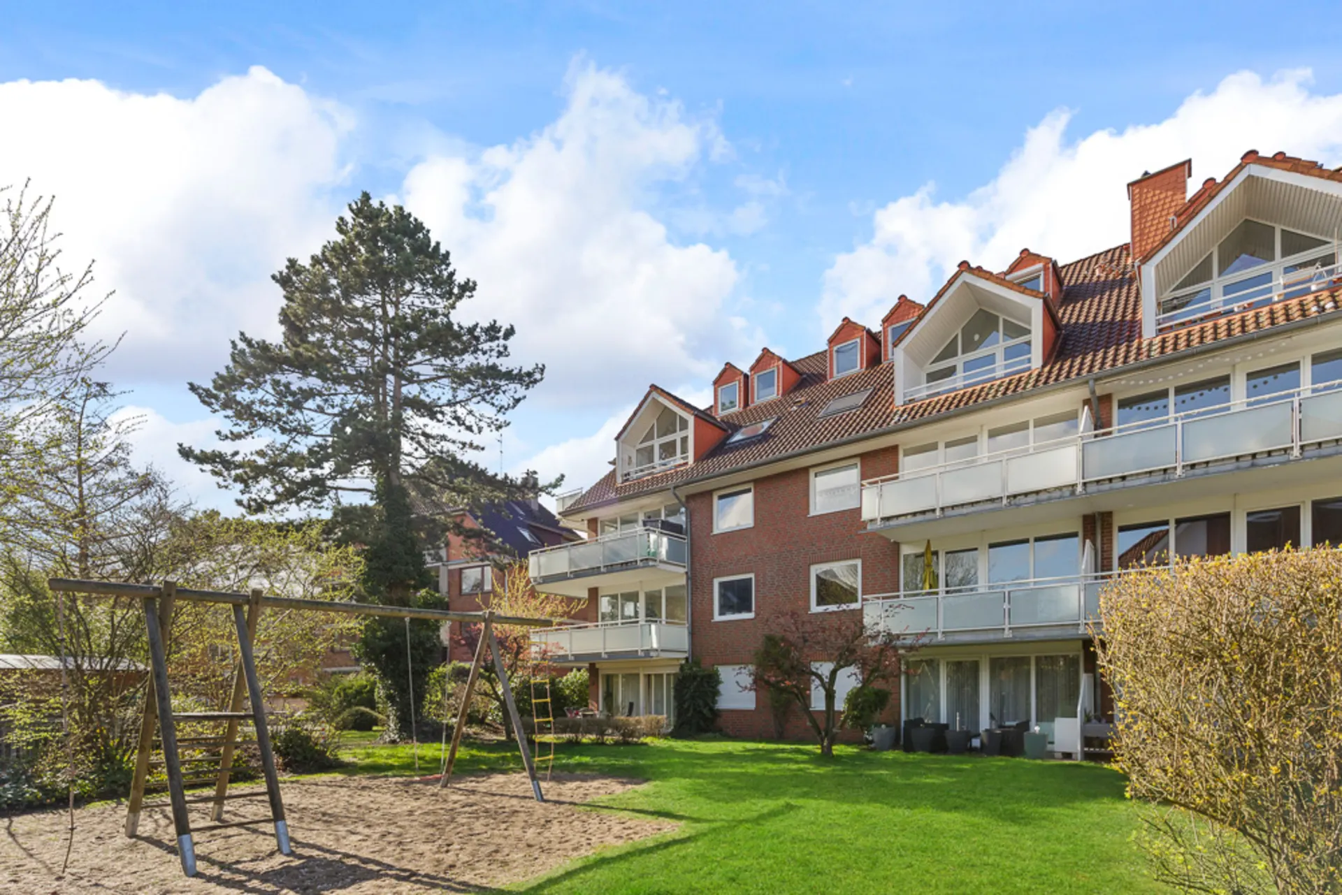 Mehrfamilienhaus mit roten Ziegeldächern, Balkonen und einem Spielplatz mit Schaukel im Garten.