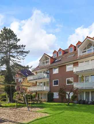 Mehrfamilienhaus mit roten Ziegeldächern, Balkonen und einem Spielplatz mit Schaukel im Garten.