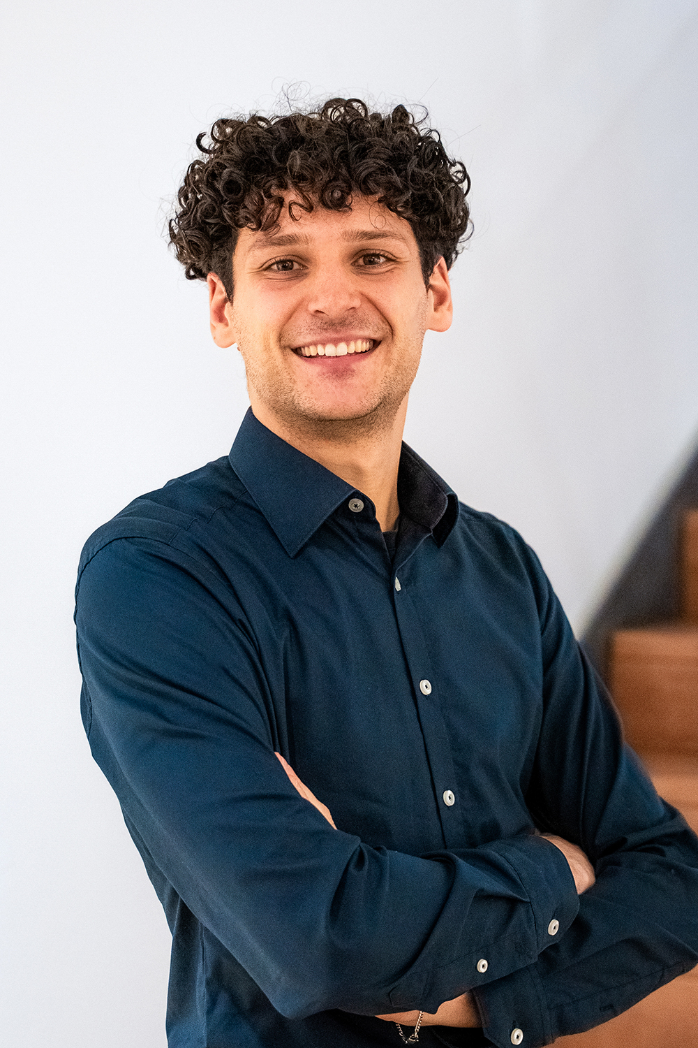 Mann mit dunklen Locken und dunkelblauem Hemd steht lächelnd vor einer weißen Wand.