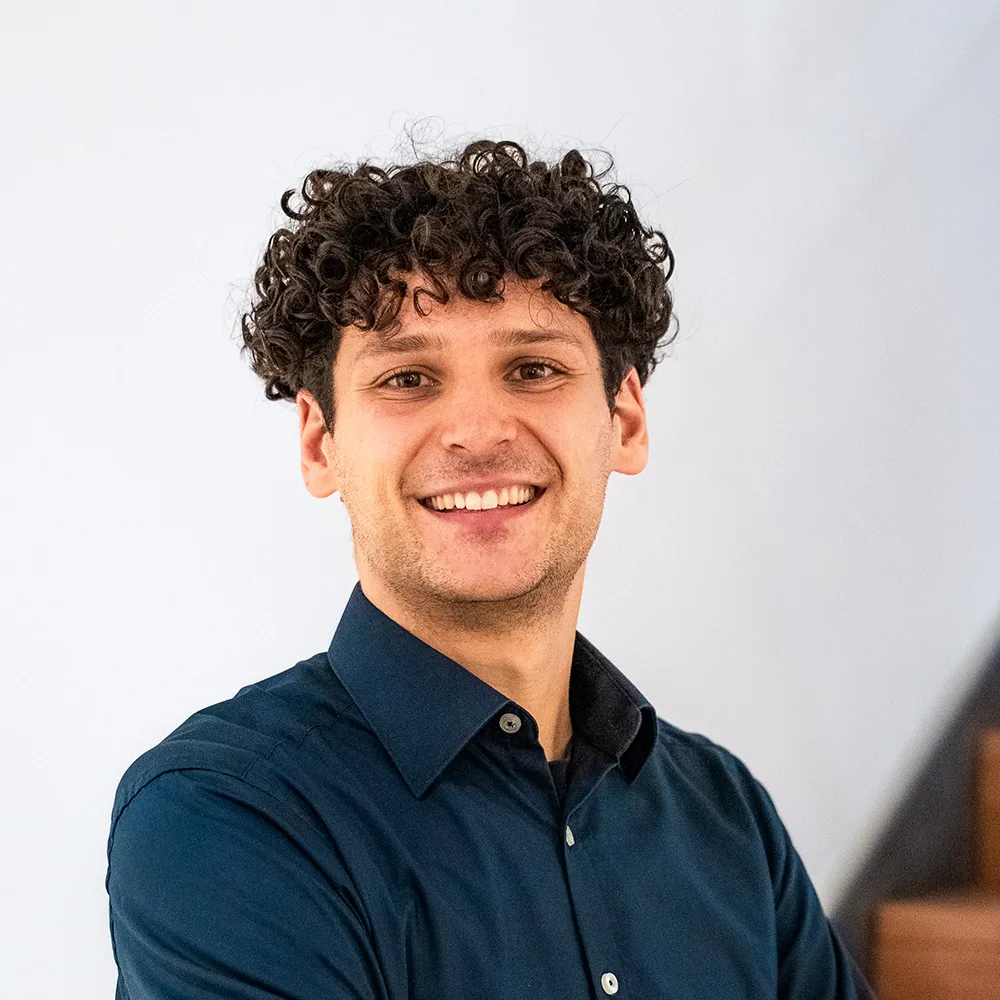 Mann mit dunklen Locken und dunkelblauem Hemd steht lächelnd vor einer weißen Wand.