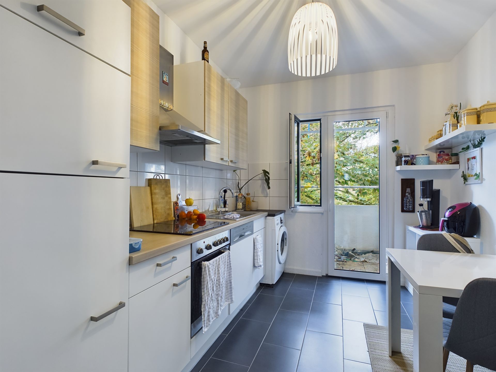 Moderne Küche mit weißen Schränken, Esstisch, Waschmaschine und Fenster zur Terrasse.