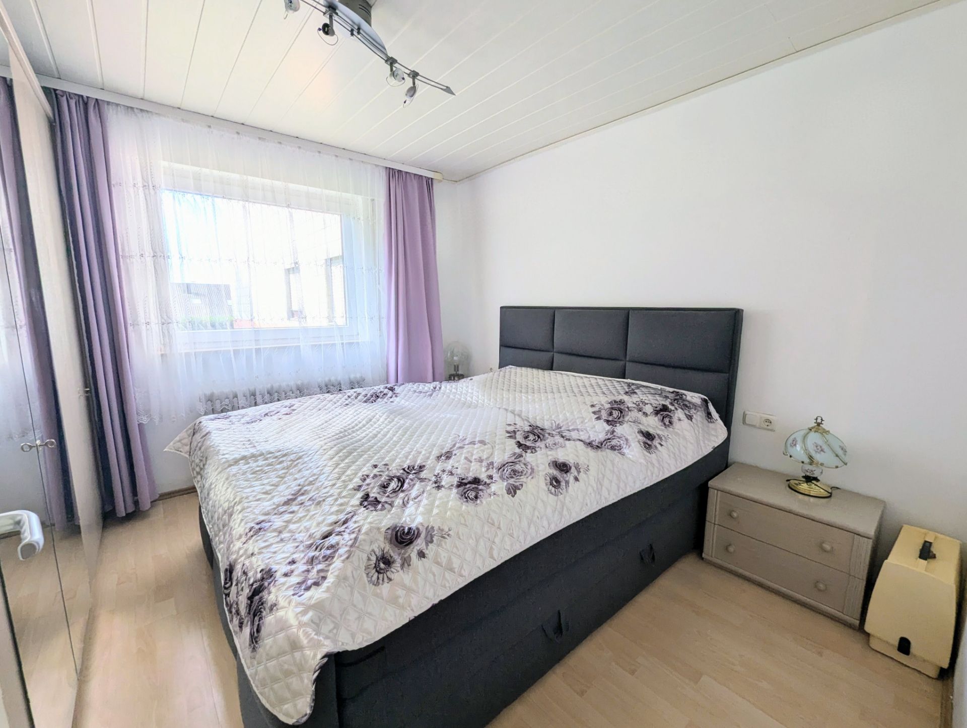 Helles Schlafzimmer mit Doppelbett, Nachttisch und Fenster mit lila Vorhängen.