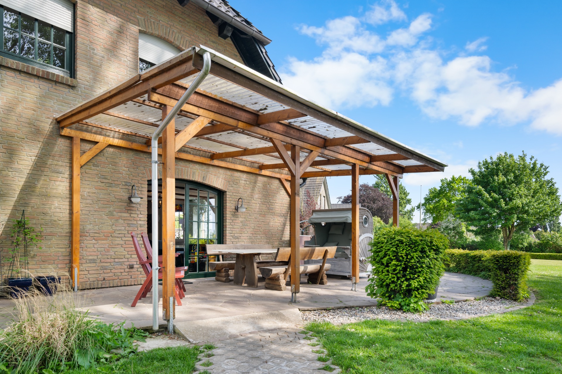 Überdachte Terrasse mit Holztisch, Bänken und Strandkorb an einem Backsteinhaus im Garten.