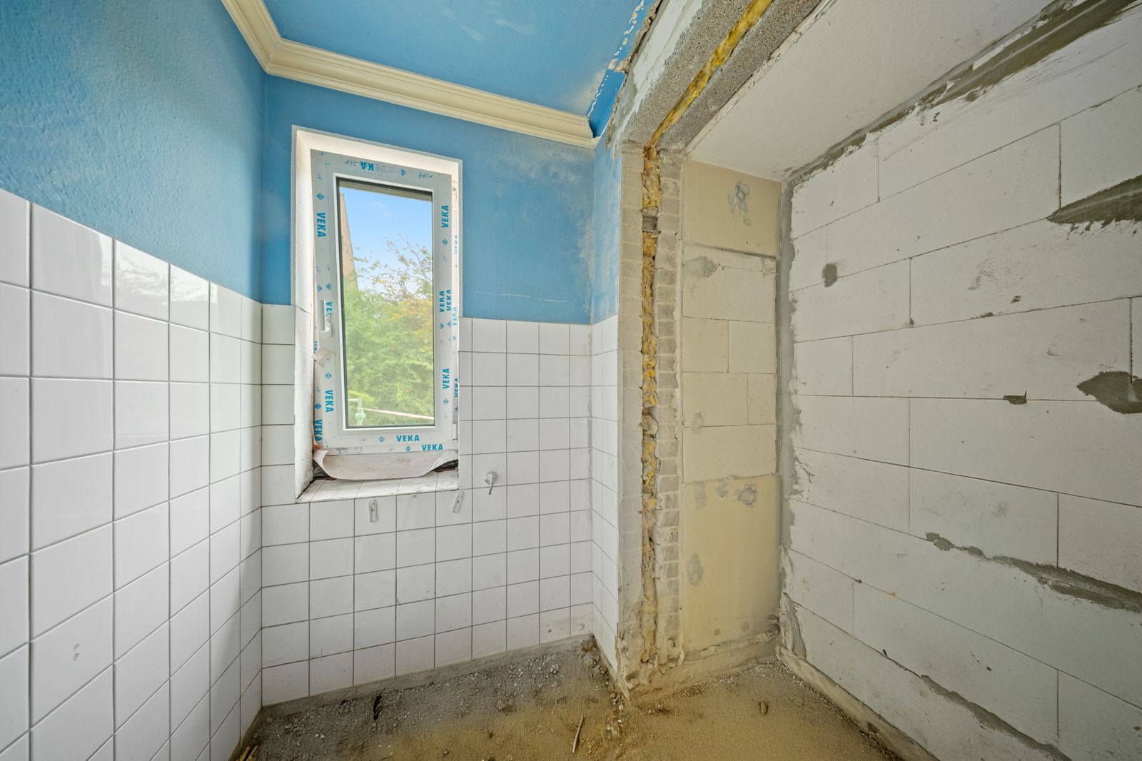 Kleiner Raum mit weißen Fliesen, blau gestrichener Wand und Fenster, teils im Umbau.