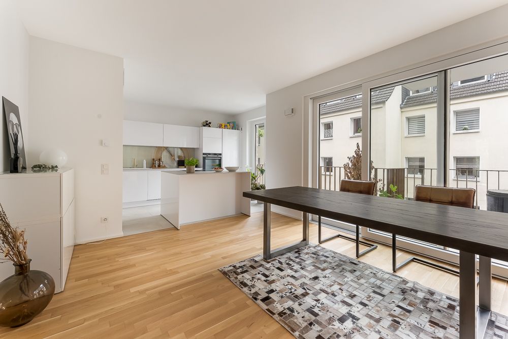 Helles Esszimmer mit Holzboden, großem Tisch und offener moderner Küche, Zugang zum Balkon.