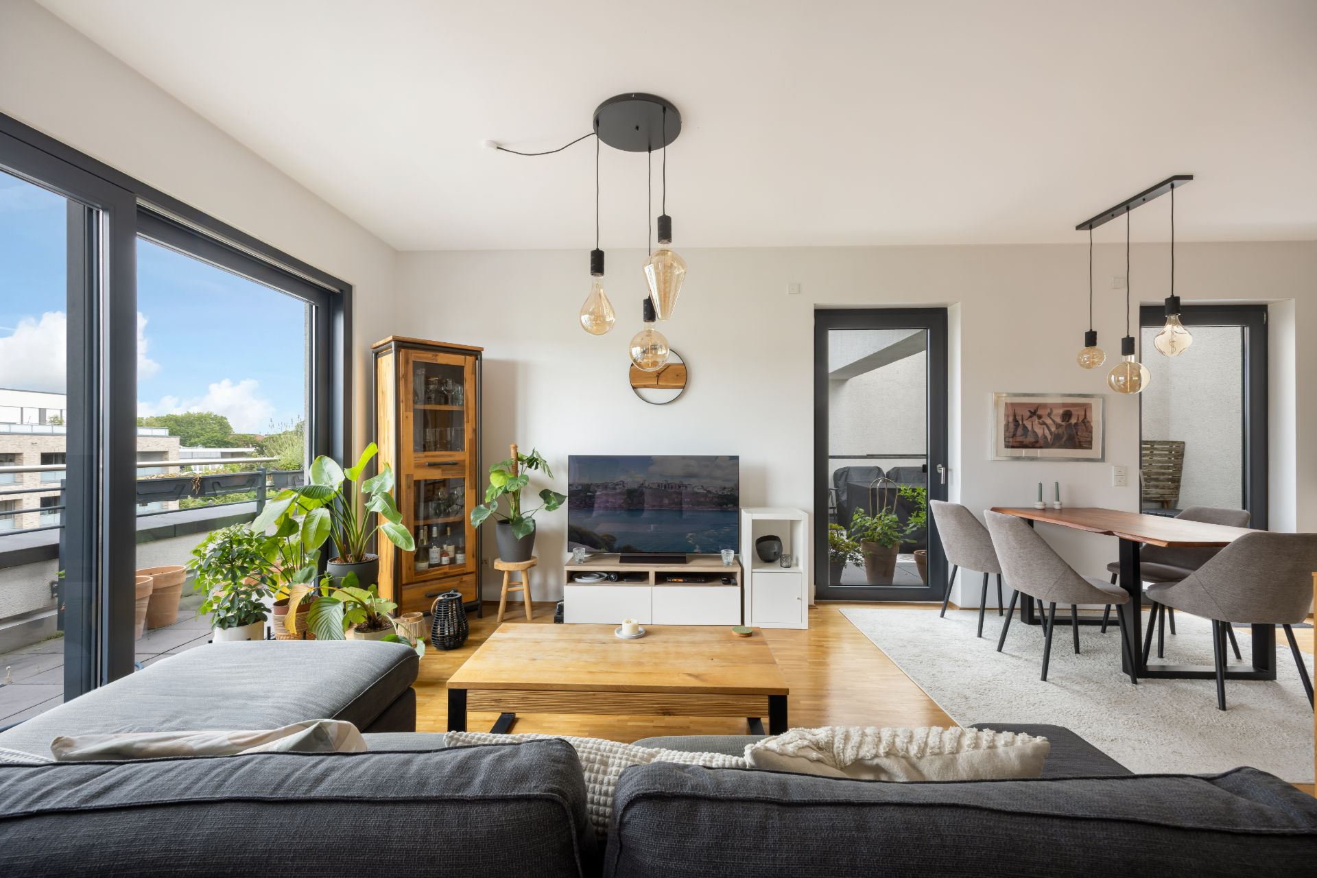 Modernes Wohnzimmer mit großen Fenstern, Sofa, Fernseher, Holztisch, Pflanzen und Essbereich.