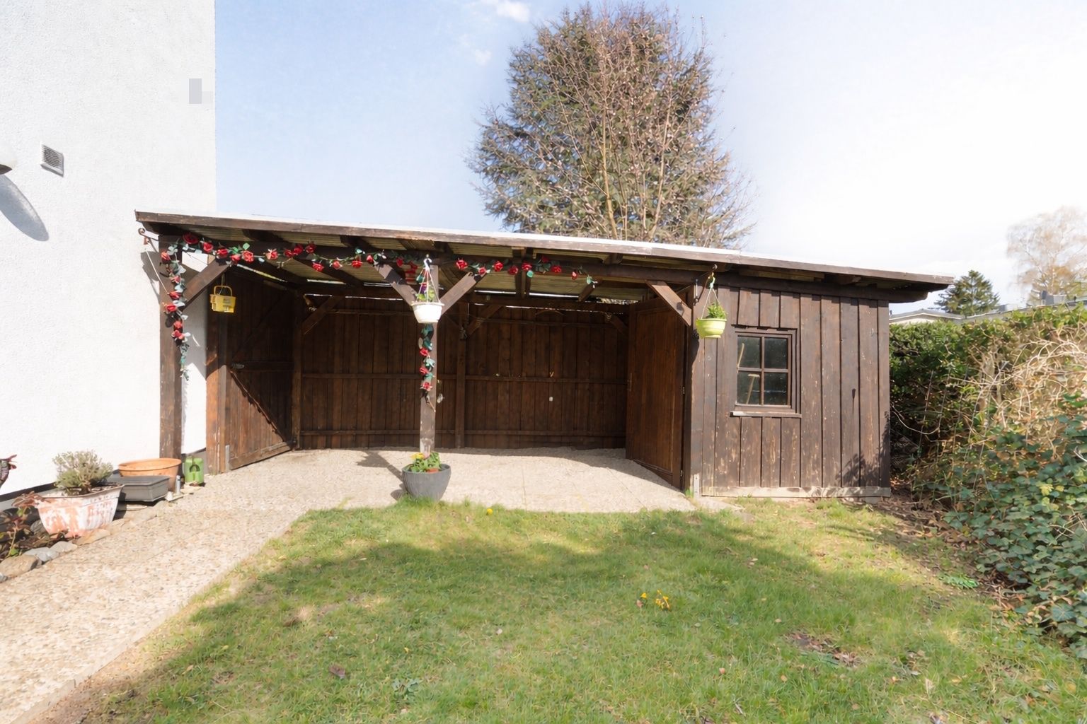 Offener Carport mit Holzwänden und Blumenampeln an einem Wohnhaus.