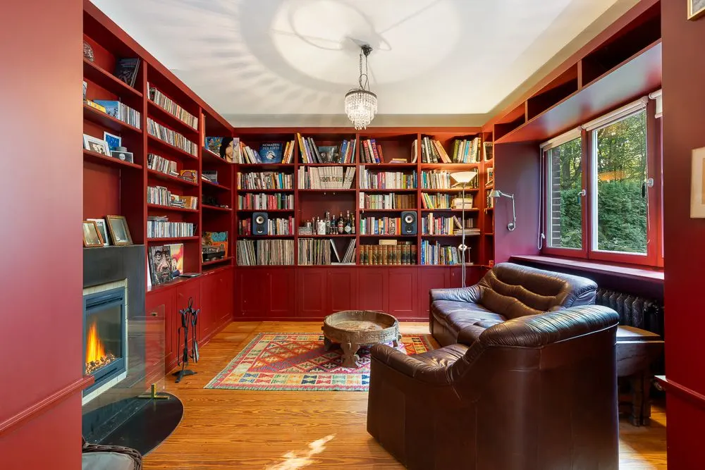 Gemütliches Wohnzimmer mit roten Bücherregalen, Ledersofa, Kamin und großem Fenster.
