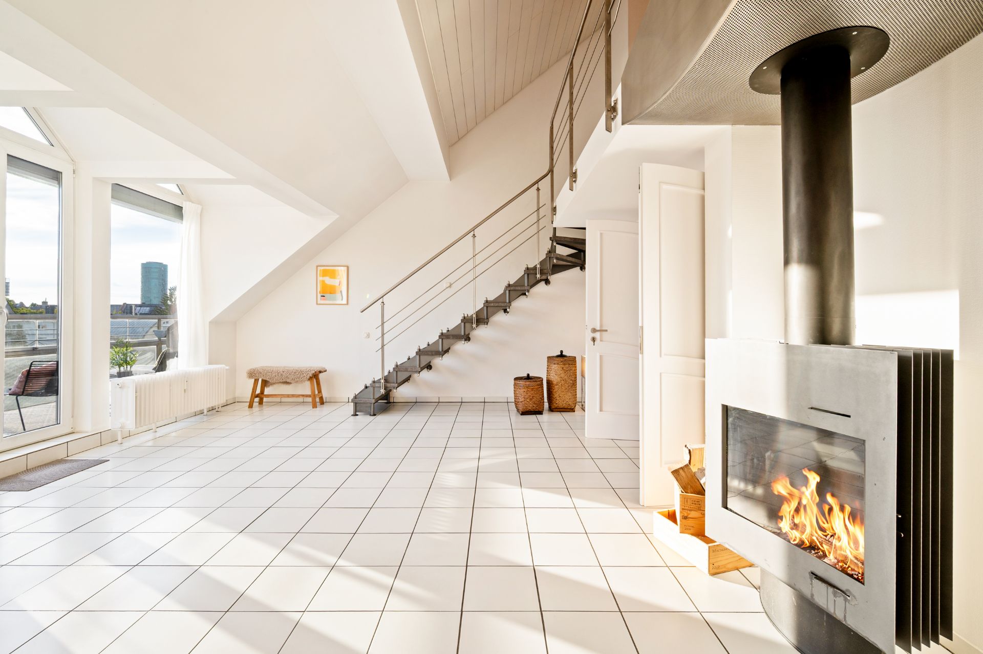 Helles Wohnzimmer mit Kamin, Fliesenboden, großer Fensterfront und Treppe ins Obergeschoss.