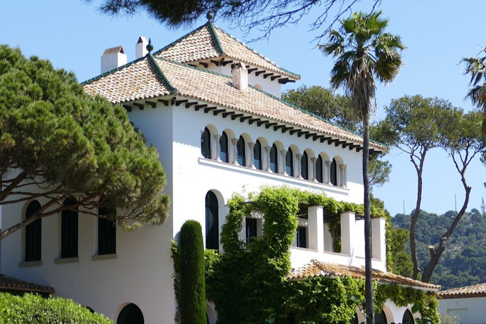 Großes weißes Wohnhaus mit grünem Dach und vielen Bögen, umgeben von Bäumen und Pflanzen.