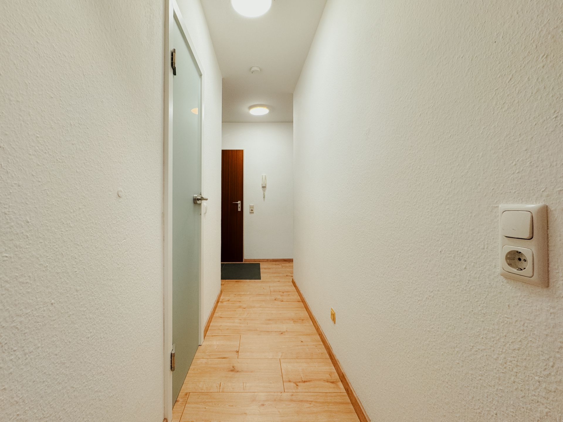 Heller Flur mit Holzboden, weiß gestrichenen Wänden und einer Eingangstür am Ende.