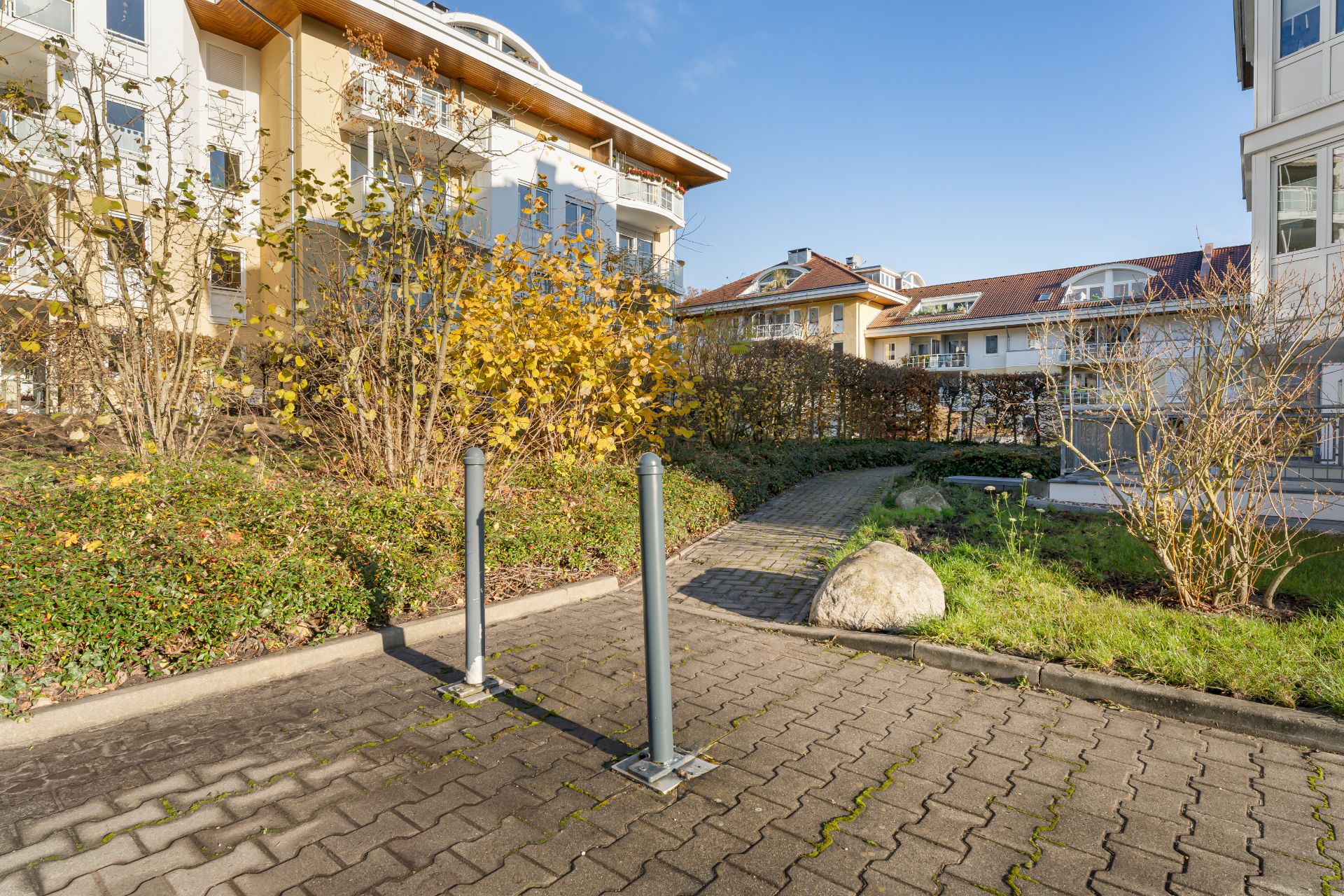 Gepflasterter Weg und Fahrradständer vor Wohngebäuden mit Büschen und Bäumen bei Sonnenschein.
