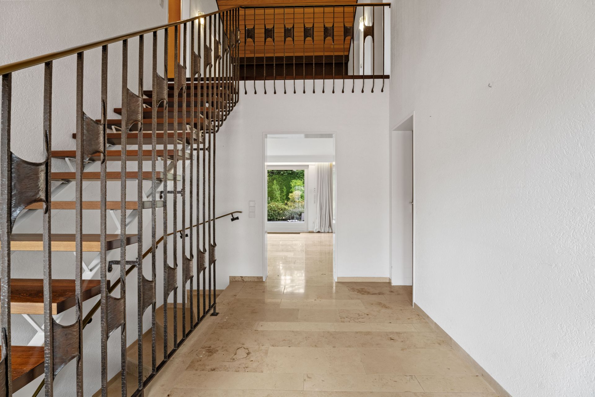 Heller Flur mit Treppe aus Holz und Metallgeländer, Blick in ein Wohnzimmer mit großem Fenster.