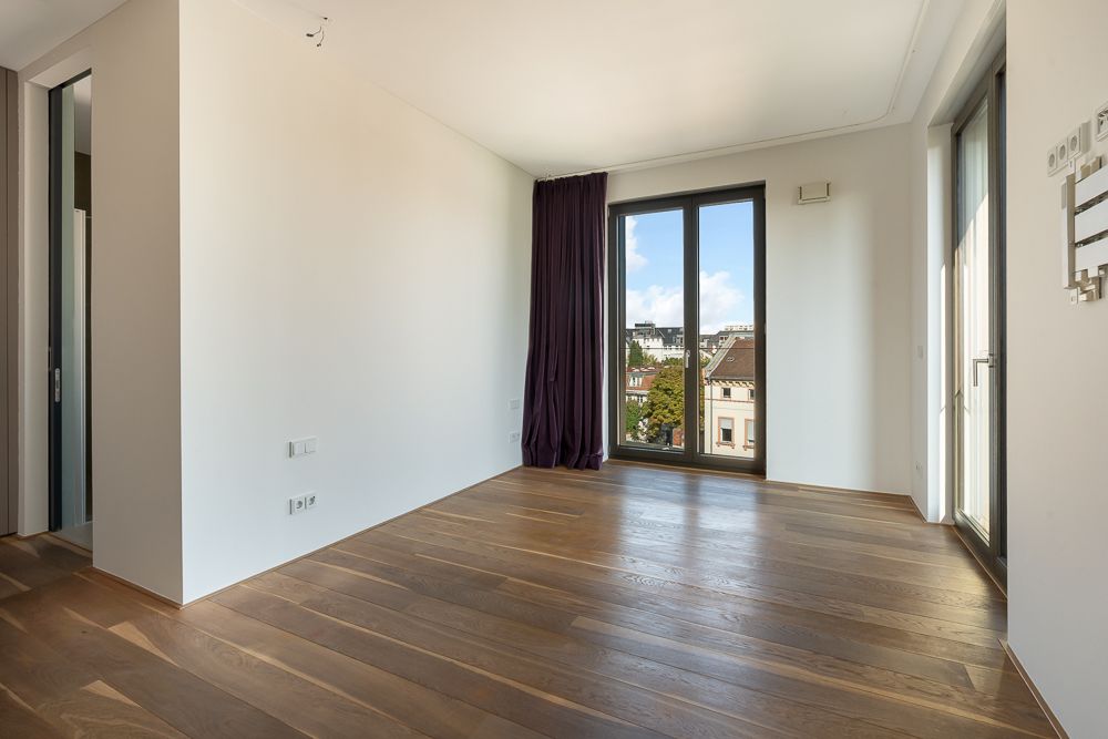 Helles Zimmer mit Holzboden, großen Fenstern und Vorhang, Blick nach draußen auf Gebäude.