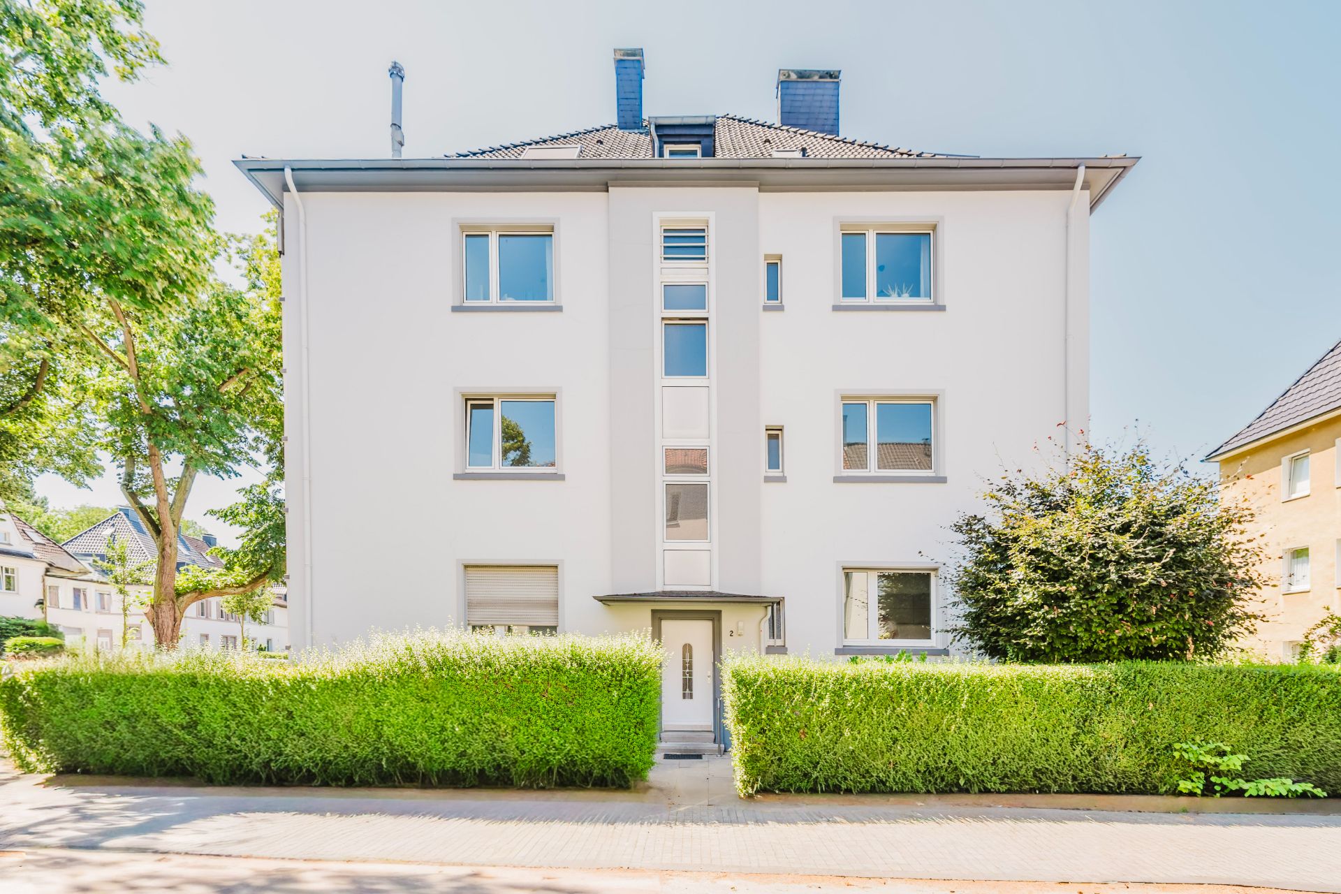Vorderansicht eines weißen, dreistöckigen Wohnhauses mit Hecke und Büsche davor.