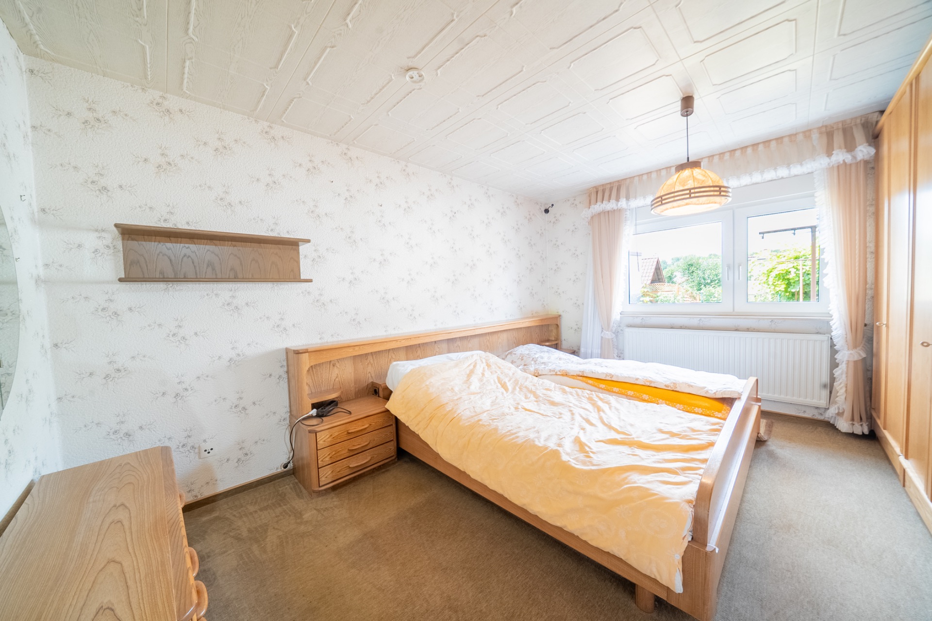 Helles Schlafzimmer mit Doppelbett, Nachttisch, Schrank und Fenster mit Vorhängen.