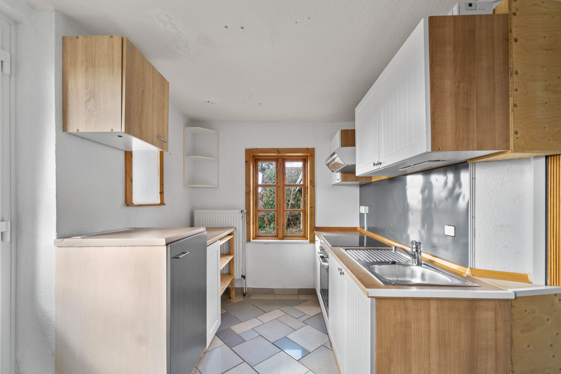 Helle Küche mit Holzschränken, Fenster, Spüle und Fliesenboden.