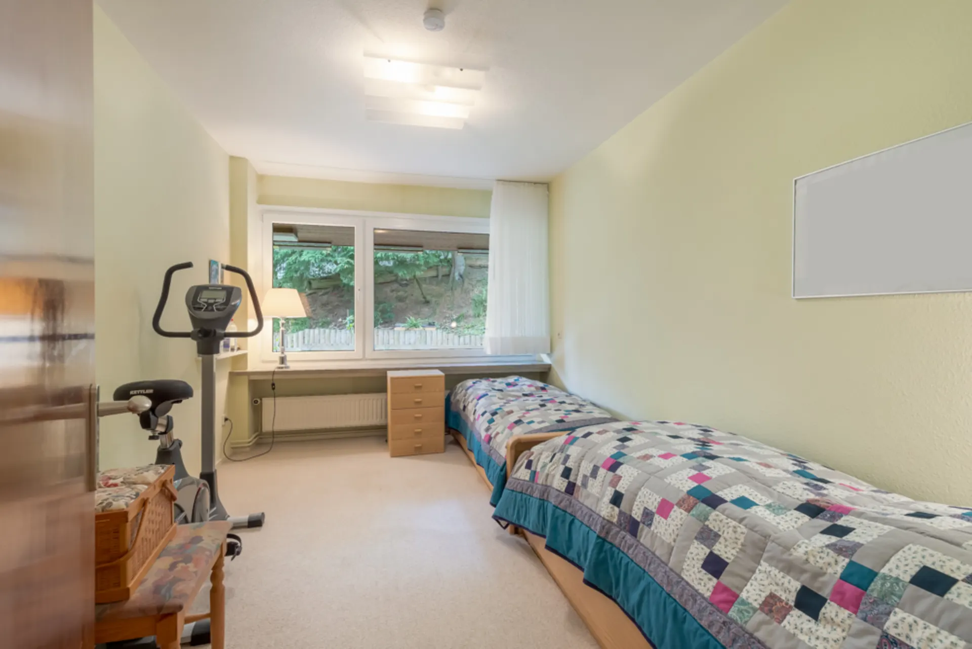 Schlafzimmer mit zwei Einzelbetten, Fenster, Schreibtisch und Heimtrainer.
