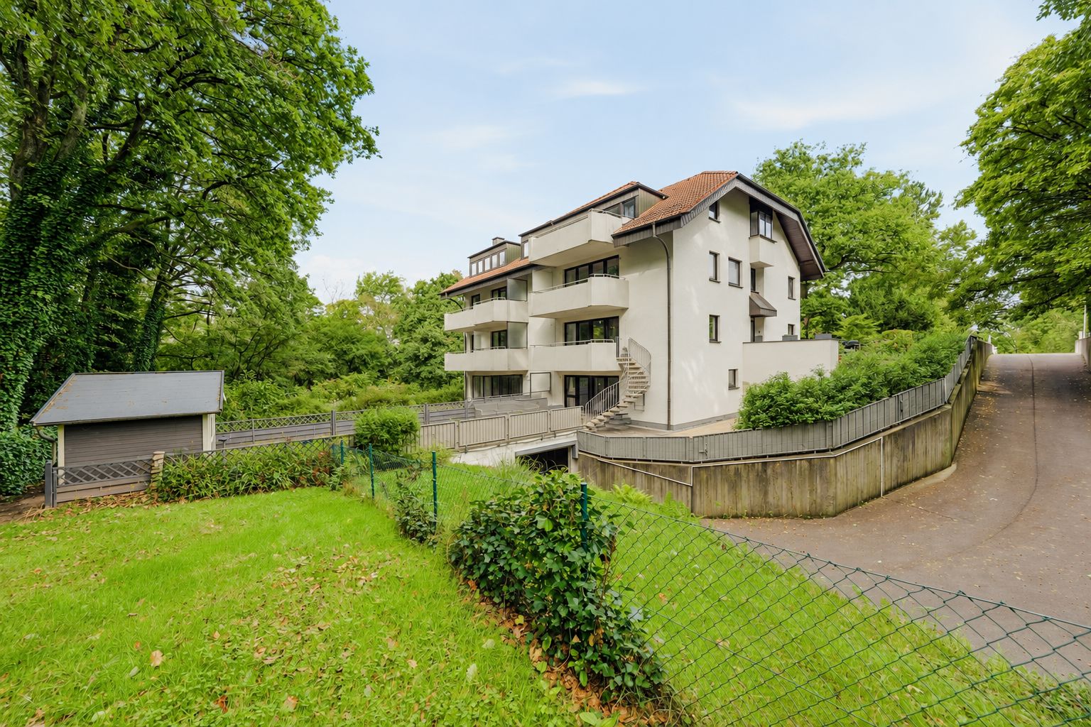 Mehrfamilienhaus mit Balkonen, umgeben von Bäumen und Garten, daneben eine kleine Hütte.