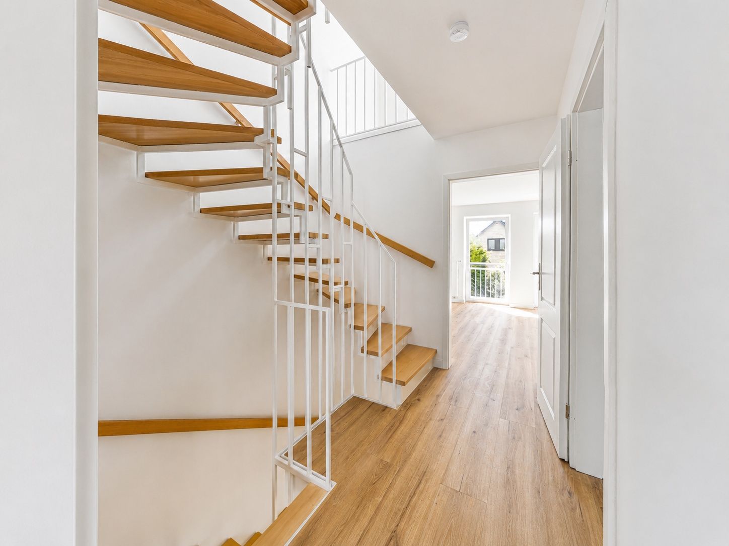 Heller Flur mit Holztreppe und offenem Zugang zu einem Raum mit großem Fenster.