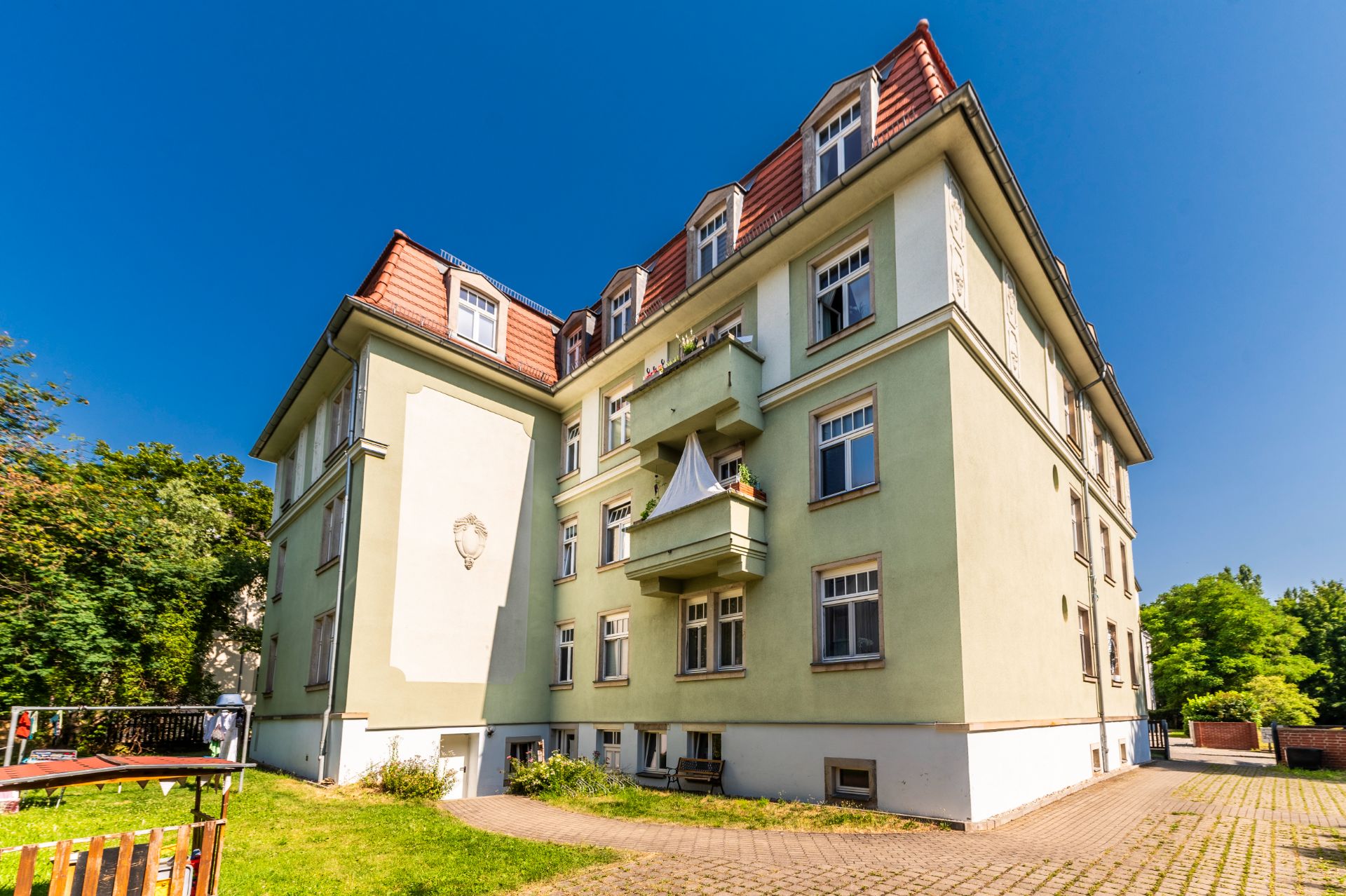 Mehrstöckiges Wohnhaus mit grünem Anstrich, Balkon und Gartenbereich bei sonnigem Wetter.