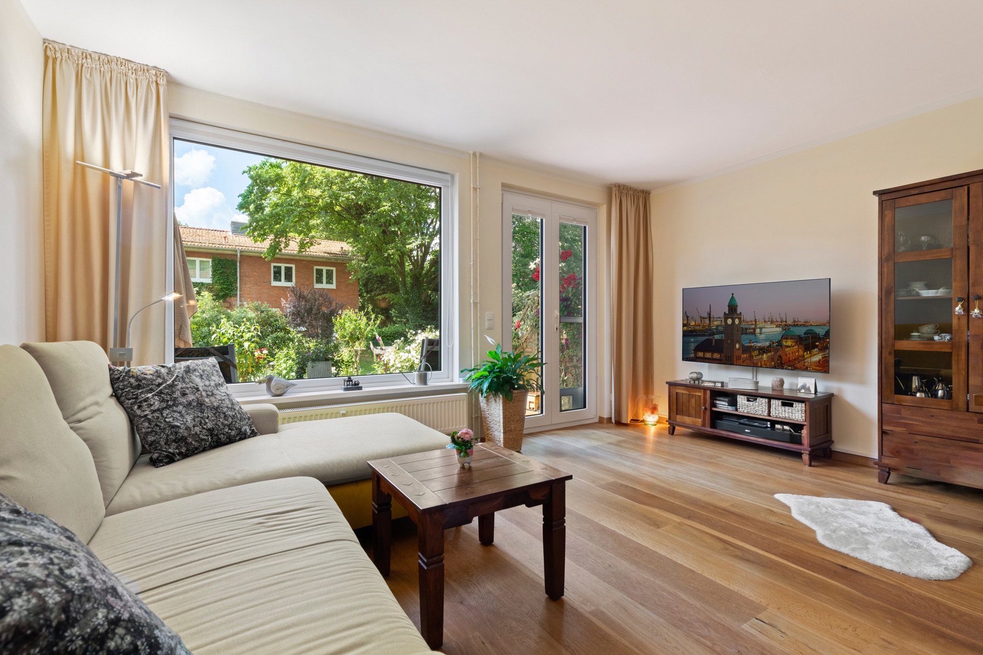 Helles Wohnzimmer mit Sofa, Holztisch, Fernseher und großem Fenster mit Gartenblick.