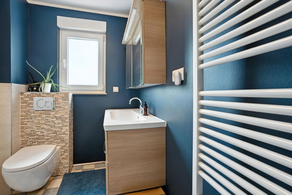 Modernes Badezimmer mit blauem Anstrich, Waschbecken, Spiegel, WC, Fenster und Handtuchheizkörper.