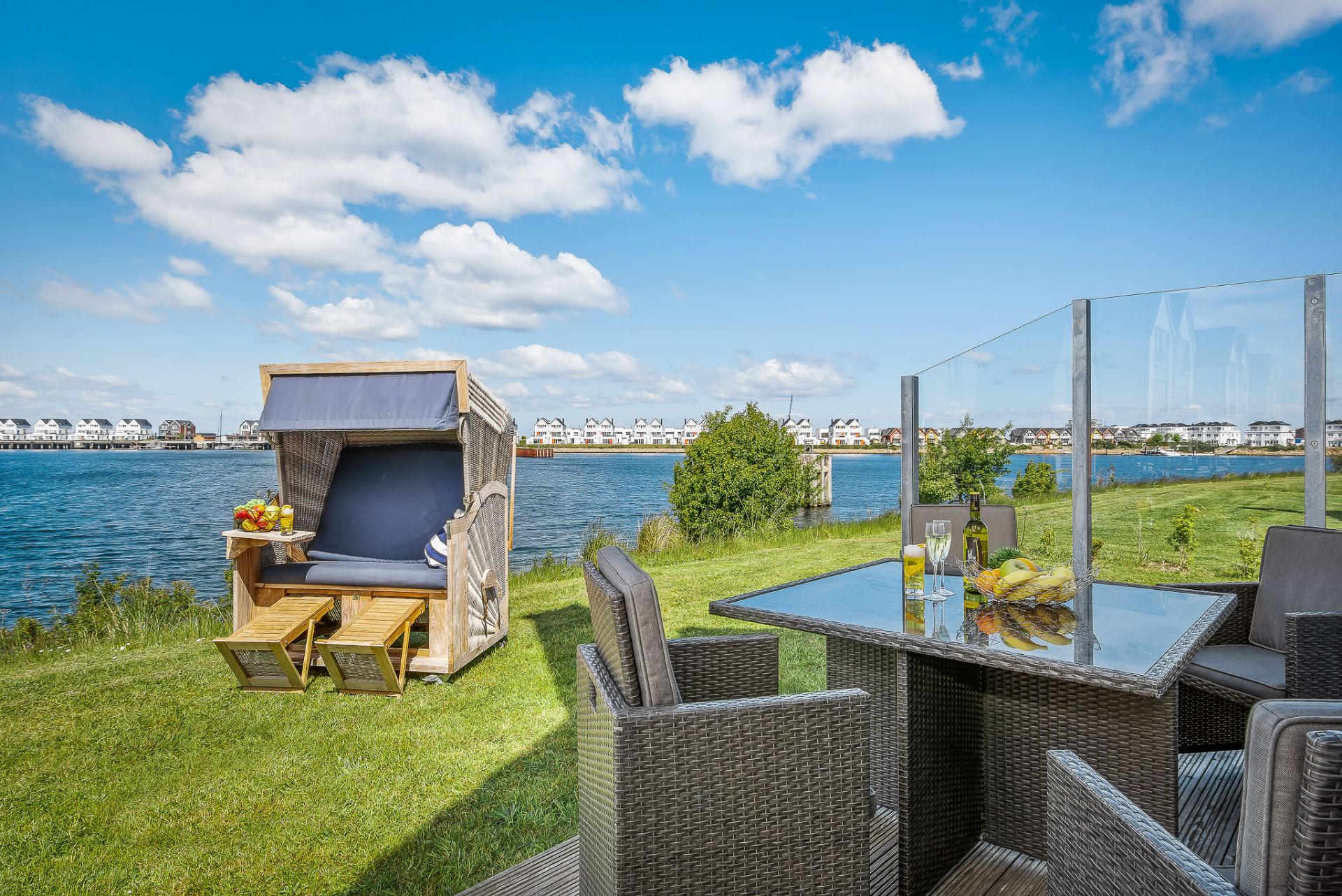 Terrasse mit Sitzgruppe und Strandkorb am Wasser, Blick auf See und Häuser im Hintergrund.