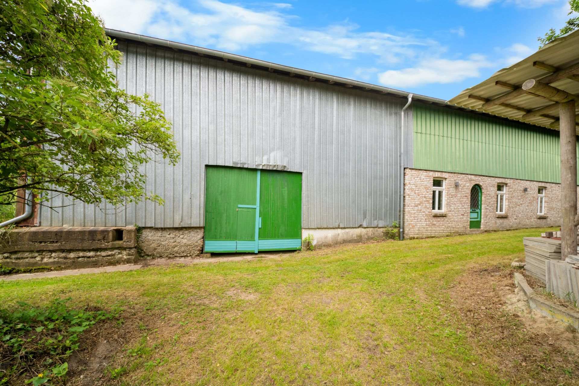 Große Halle aus Blech und Backstein mit grünen Türen und Fenstern, davor Rasenfläche.