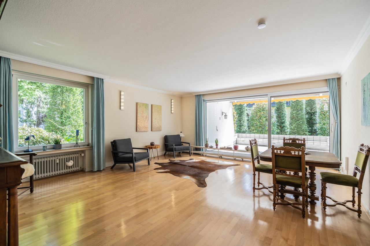 Helles Wohnzimmer mit großen Fenstern, Holzboden, Sitzecke und Esstisch mit Stühlen.