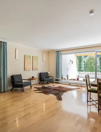 Helles Wohnzimmer mit großen Fenstern, Holzboden, Sitzecke und Esstisch mit Stühlen.