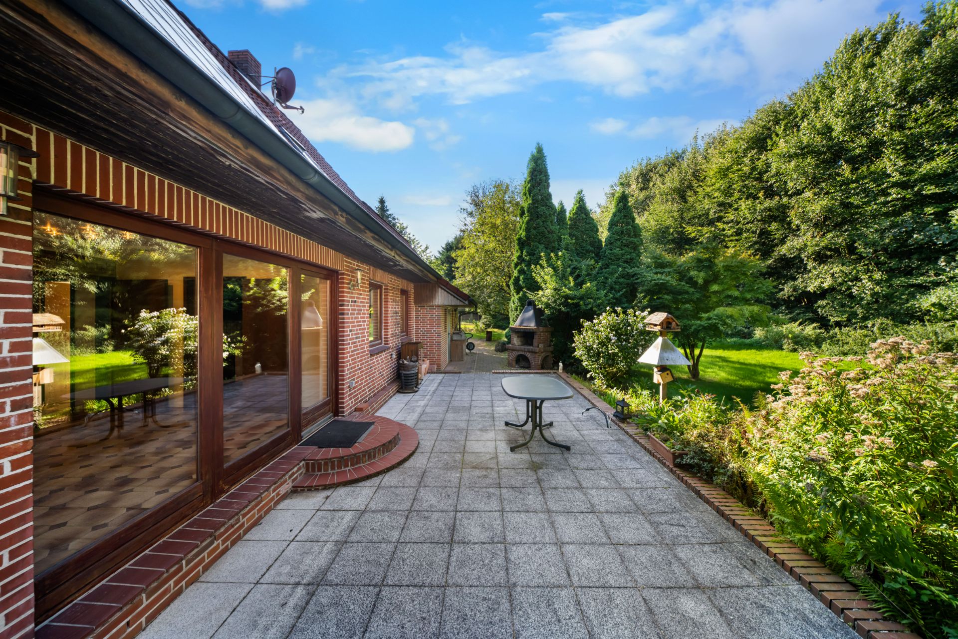 Große Terrasse mit Glastür, rundem Tisch, Grill und Blick auf Garten mit Büschen und Bäumen.