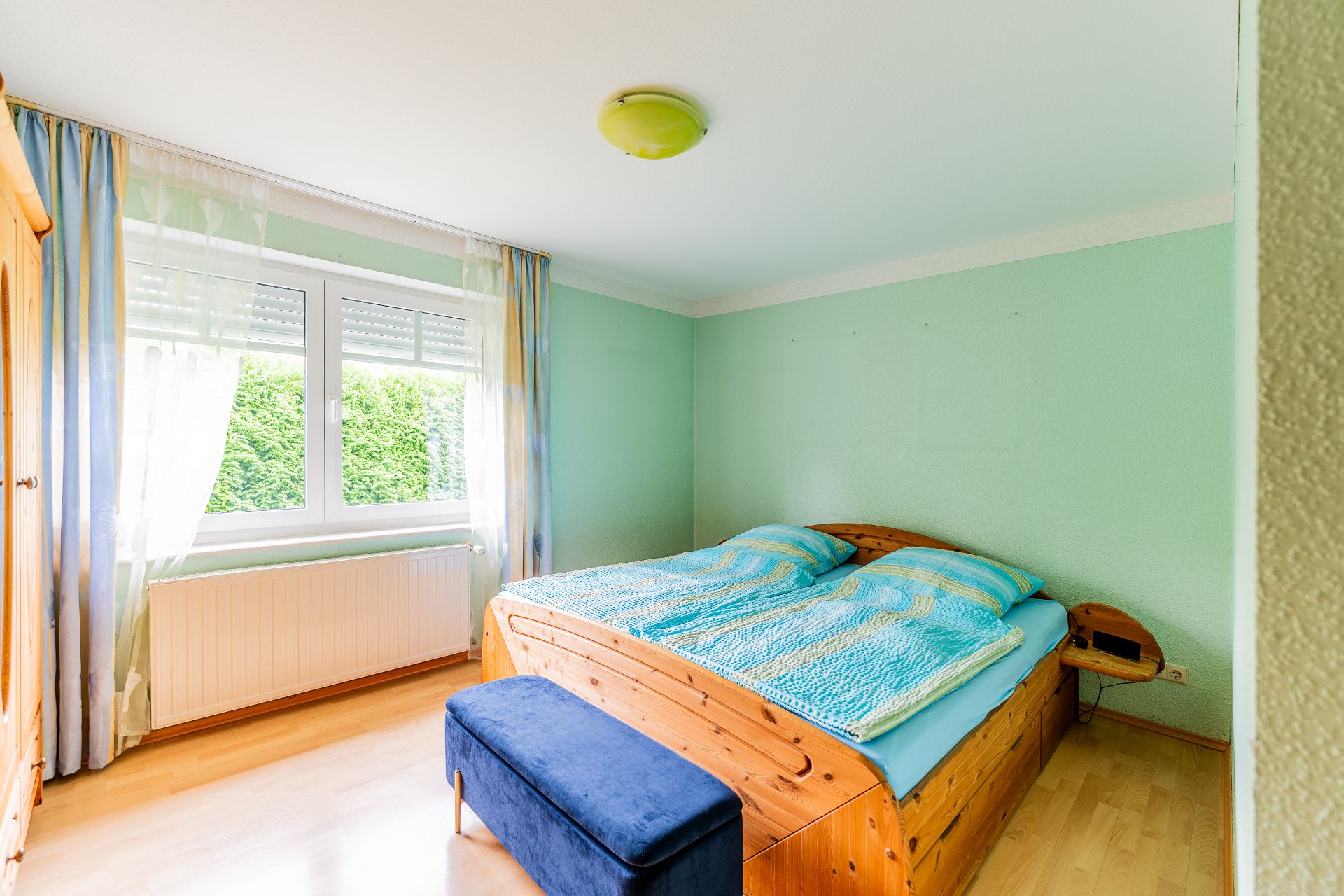 Helles Schlafzimmer mit Doppelbett, großem Fenster, blauen Gardinen und Holzfußboden.