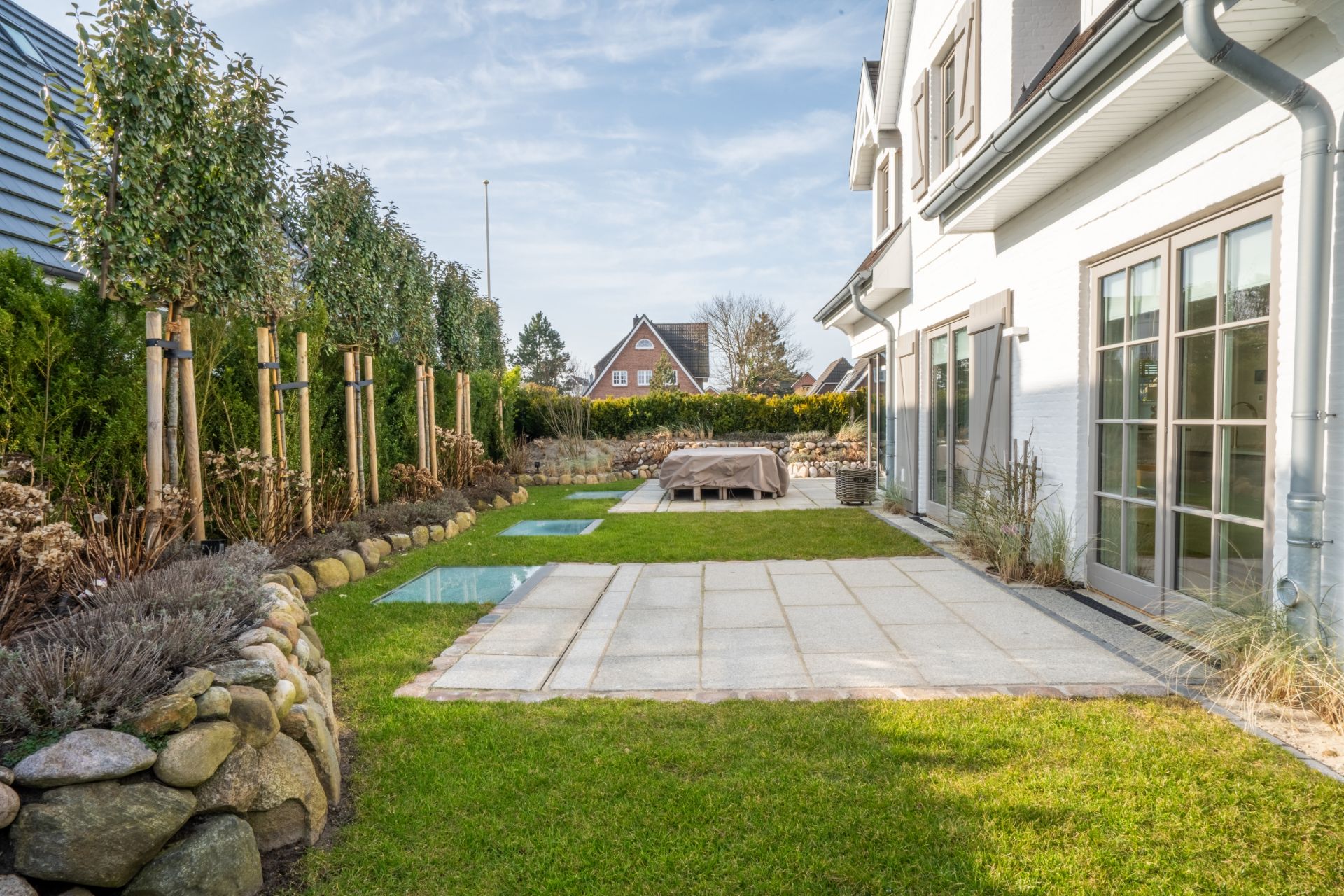 Garten mit gepflasterter Terrasse, Rasenfläche und einigen Bäumen neben einem Wohnhaus.